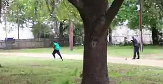 Video image of Scott in a turquoise shirt and black pants running away to the left, many meters away, with Slager in uniform pointing his gun at Scott in the right of the screen