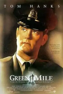 The words Tom Hanks, a correctional officer looking to the distance, below the words The Green Mile, in the middle of the words, a small silhouette of a big man and a small man walking towards a light.